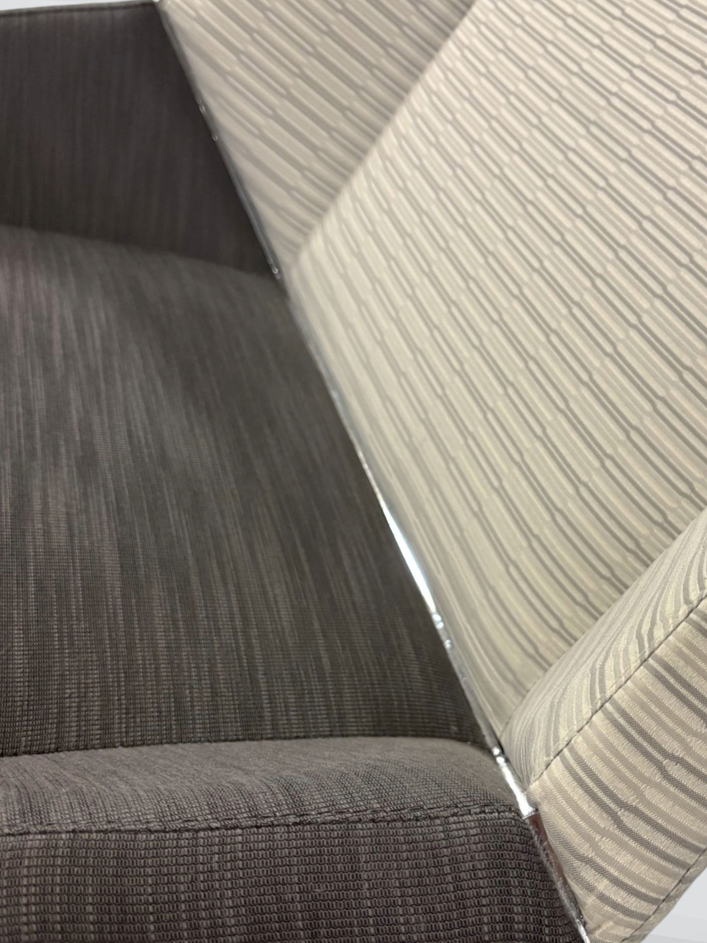 Close-up of distinctive metal accent detail on a modern upholstered chair or sofa corner, showing dark gray textured seat fabric and a light beige striped backrest.