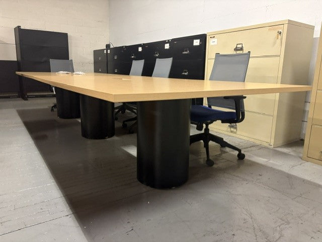A long rectangular office conference table with a maple wood top and black cylindrical pedestal bases, shown in a warehouse or office setting with office chairs, filing cabinets, and a beige cabinet in the background.