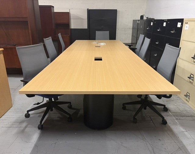 A Maple wood conference table with a black cylindrical pedestal base, surrounded by gray mesh office chairs in a warehouse showroom setting.