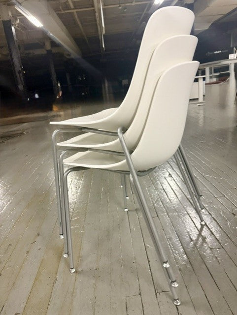 Three white molded plastic dining chairs with chrome metal legs stacked together on a polished wood floor in an indoor showroom.