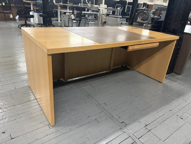 A light wood office desk with a single drawer and a recessed center panel, photographed in a warehouse or office space with gray floorboards and other furniture in the background.