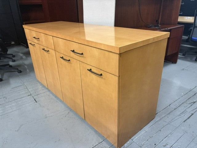 A light wood office credenza or sideboard cabinet with three drawers on the top and three cabinet doors below, shown in an office or warehouse setting on a concrete floor.