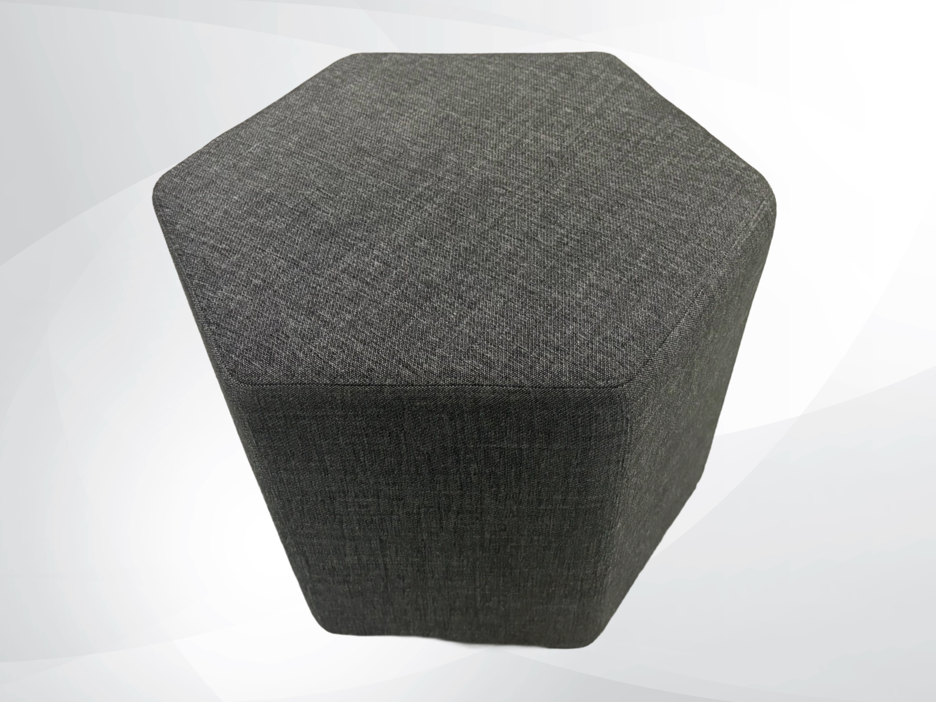 A dark gray hexagonal upholstered ottoman with a textured fabric cover, shown against a light gray and white abstract background.