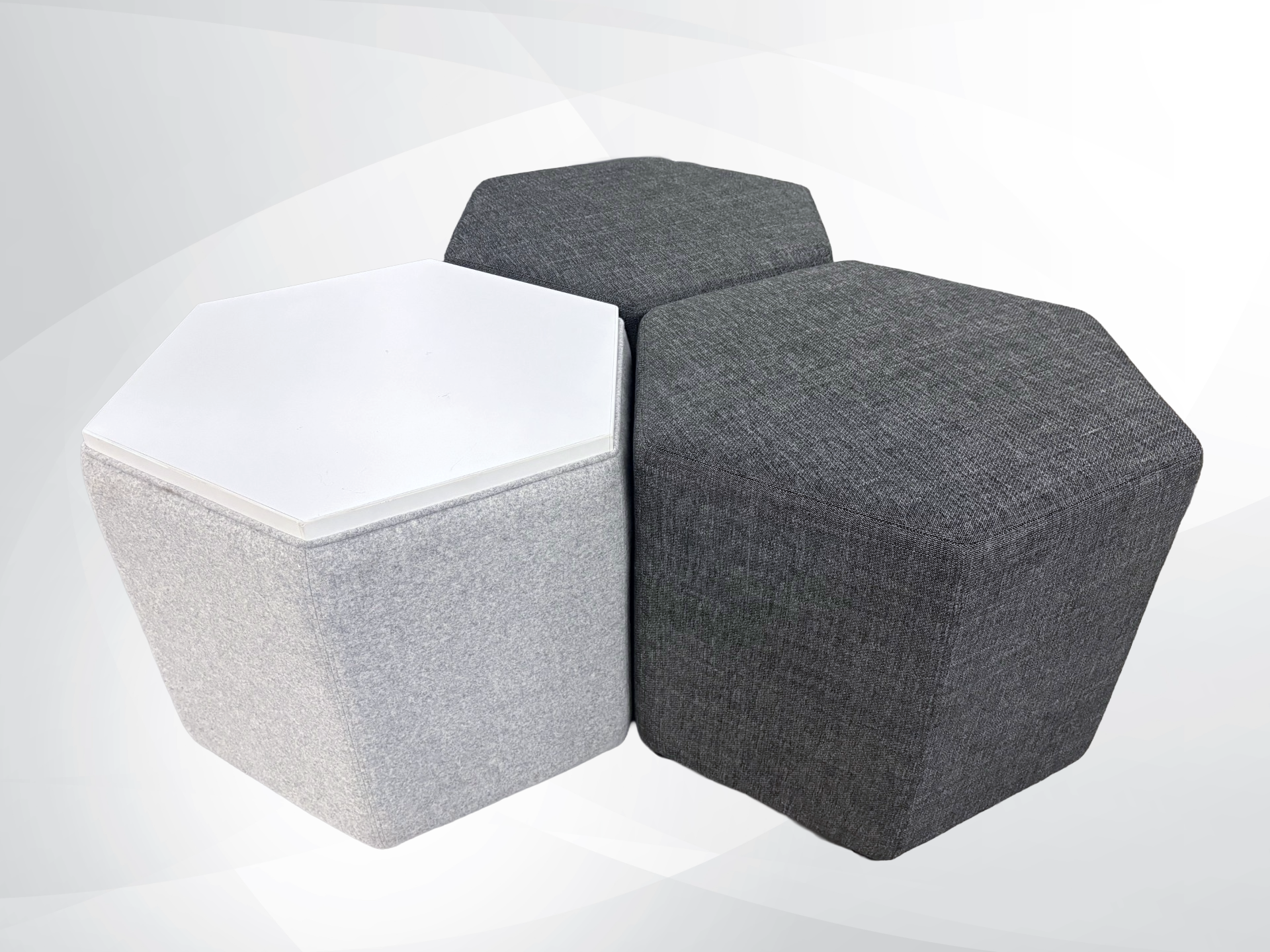Three modern hexagonal upholstered ottomans or small tables arranged together on a light gray studio background. One ottoman has a white top and light gray fabric sides, while the other two have dark gray fabric tops and sides.