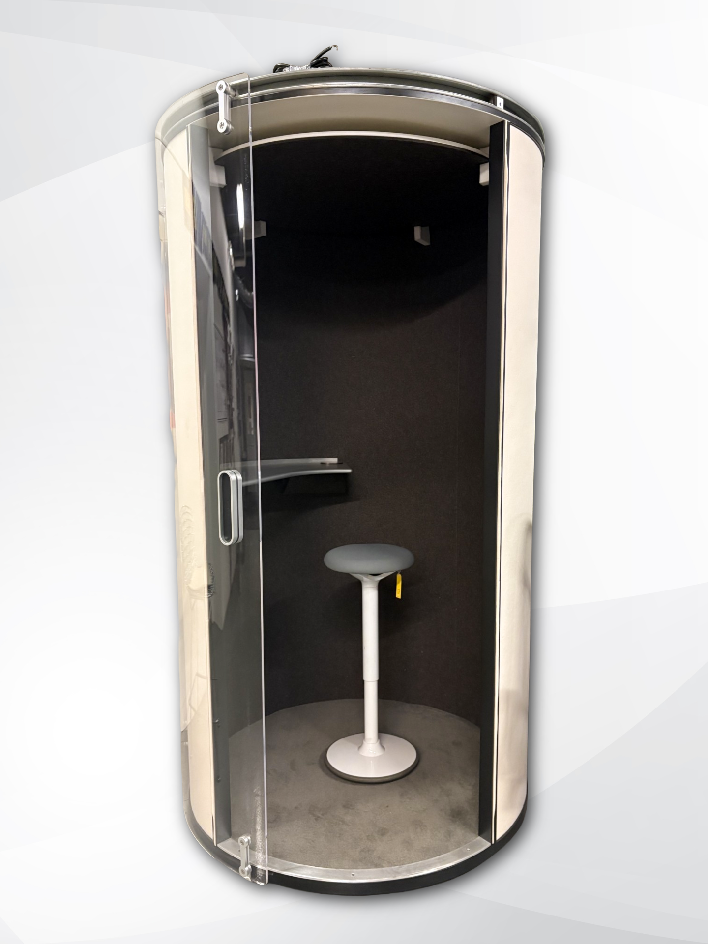 A modern, cylindrical soundproof phone booth with a clear glass door, dark interior, and a small white stool in the center, shown against a light gray background.