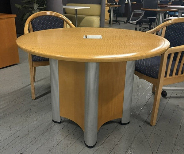 An oval wood conference table with a light oak finish and silver pedestal legs, shown with two matching wooden chairs featuring blue patterned upholstery in an office setting.