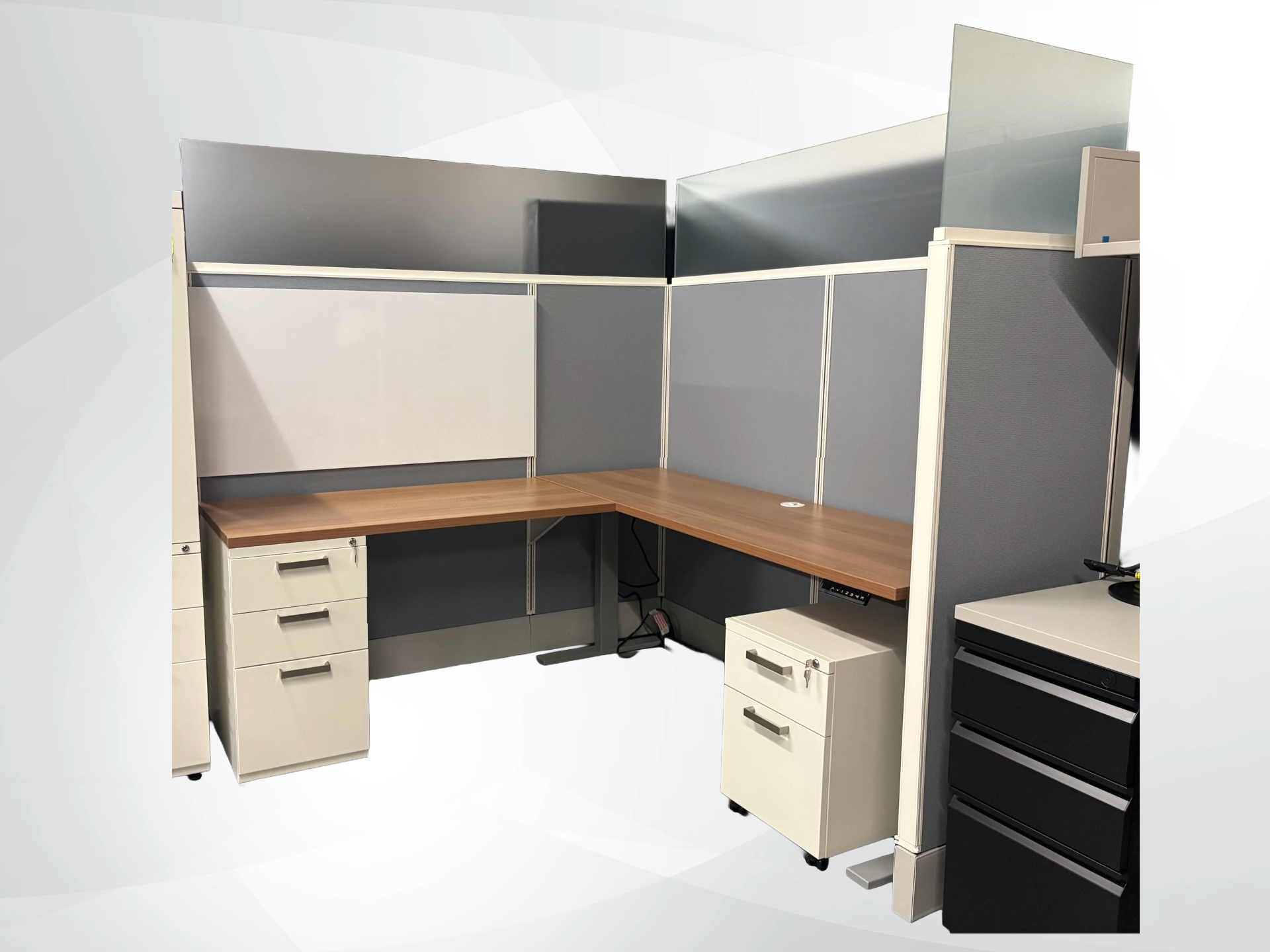 L-shaped office cubicle desk setup with gray and white panels, wood-look desktop surfaces, cream-colored filing cabinets, and a black drawer unit, shown on a light gray studio background.