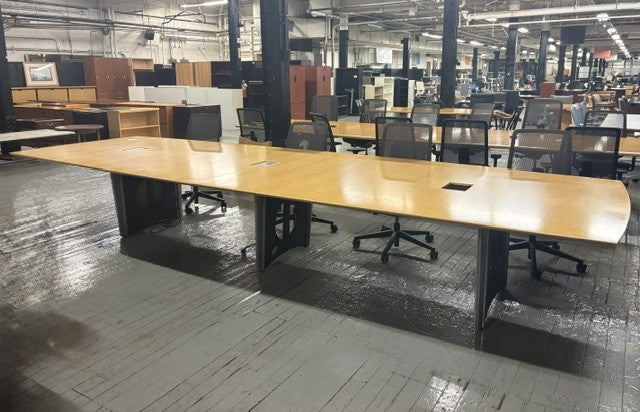 A large rectangular office conference table with a light wood top and black metal legs, shown in a warehouse or showroom with other desks and chairs around it.