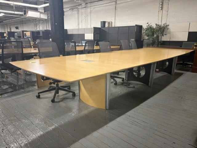 A large rectangular office conference table with a light wood grain top and curved silver metal base, surrounded by gray mesh office chairs in an office space.
