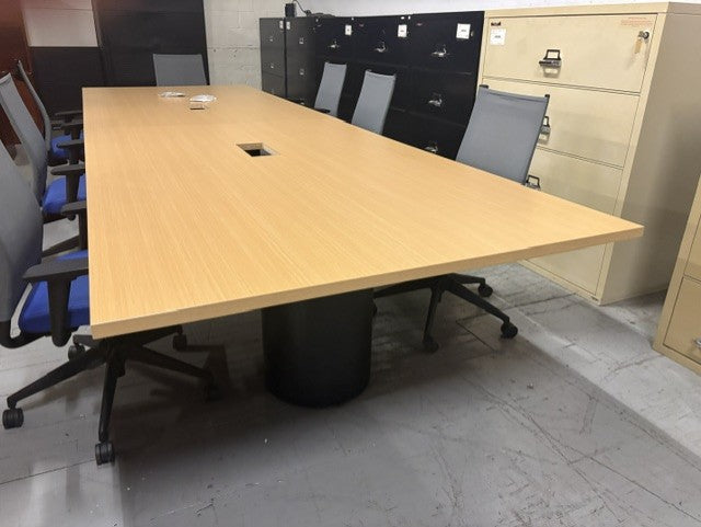 A large rectangular meeting table with a Maple top and a black pedestal base, surrounded by gray office chairs in a warehouse or office setting.