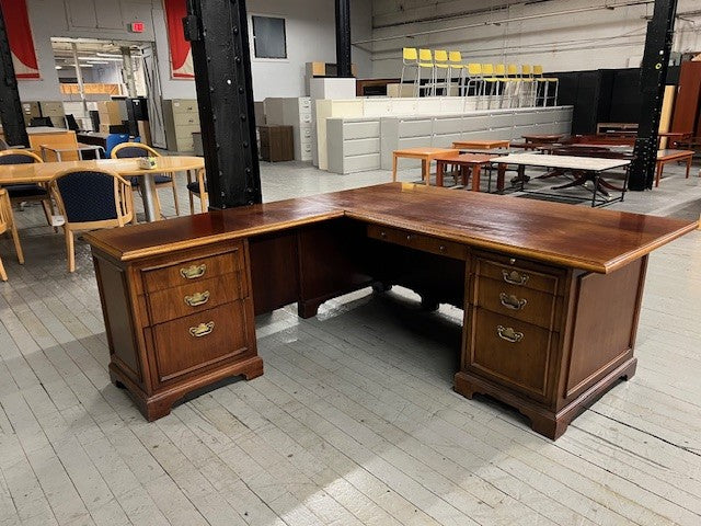 Wooden L-shaped desk in a warehouse setting with other furniture and equipment.